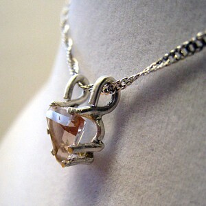 May include: A silver-toned necklace featuring a heart-shaped, peach-colored gemstone pendant. The pendant is held in place by a decorative silver setting and hangs from a delicate chain. The necklace is displayed against a neutral background.
