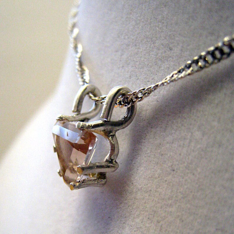 May include: A silver-toned necklace featuring a heart-shaped, peach-colored gemstone pendant. The pendant is held in place by a decorative silver setting and hangs from a delicate chain. The necklace is displayed against a neutral background.
