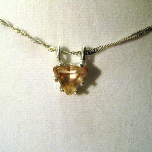 May include: A silver necklace featuring a triangular, light orange gemstone pendant. The pendant is held by silver prongs and hangs from a delicate silver chain. The gemstone has a faceted surface, catching the light.