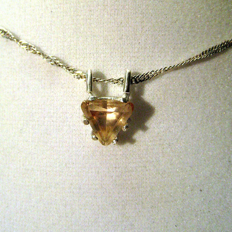 May include: A silver necklace featuring a triangular, light orange gemstone pendant. The pendant is held by silver prongs and hangs from a delicate silver chain. The gemstone has a faceted surface, catching the light.
