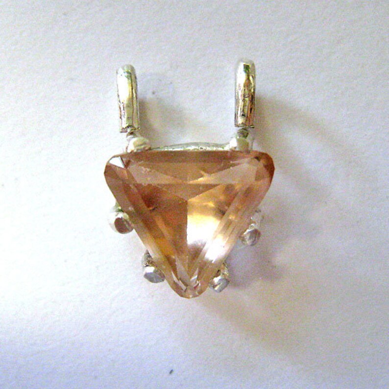 May include: A pendant featuring a large, triangular, faceted, amber-colored gemstone set in a silver-toned metal frame. The pendant has two small loops at the top for a chain and small decorative prongs around the gemstone.