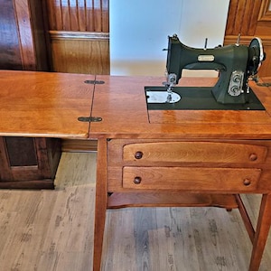 May include: A vintage sewing machine on a wooden table. The machine is dark green and black, with a silver needle and presser foot. The table is made of light brown wood and has two drawers. The table's side panels fold out for extra workspace.