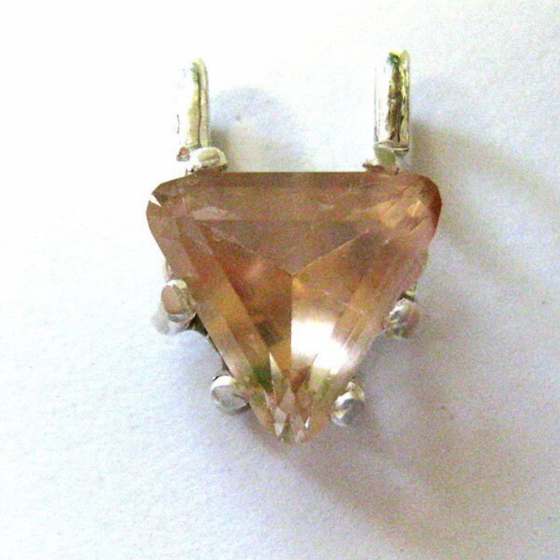 May include: A triangular pendant with a faceted, peach-colored gemstone. The gemstone is set in a silver-colored metal setting with two loops at the top for a chain. The pendant has a pointed bottom and is displayed against a white background.