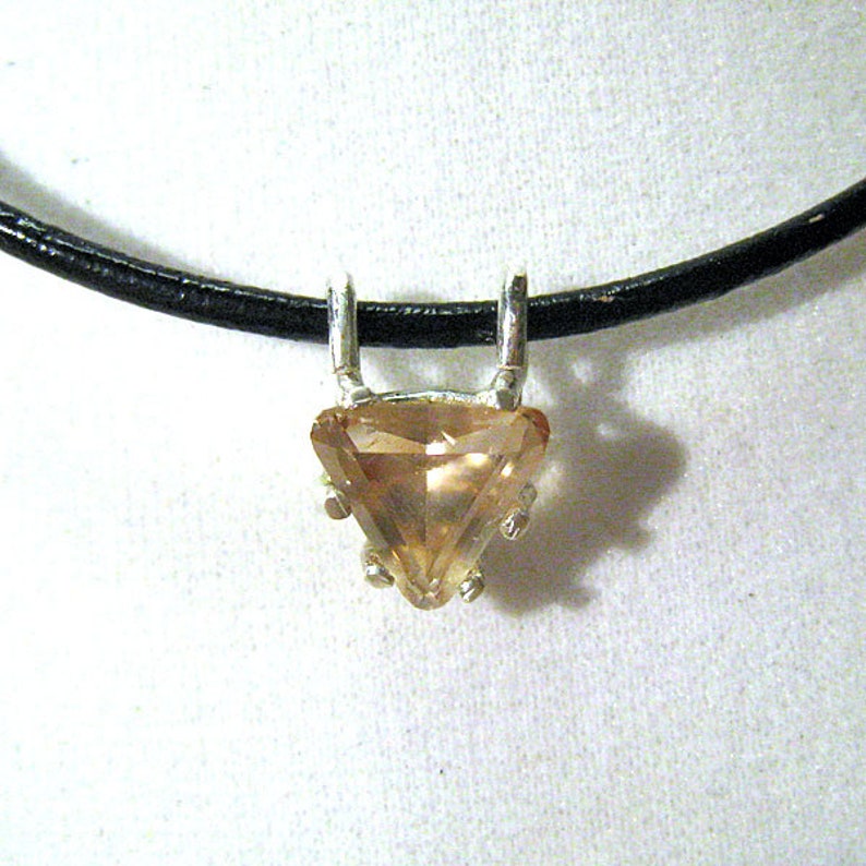 May include: A pendant necklace featuring a triangular, light brown gemstone set in a silver-colored metal. The pendant hangs from a black cord, creating a simple yet elegant design. The gemstone has multiple facets, reflecting light.