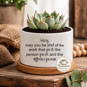 May include: A white ceramic cylindrical planter containing a green succulent plant. The planter features black printed text that reads: Stacey, may you be proud of the work that you do the person you are and the difference you make. It sits on a round wooden base.