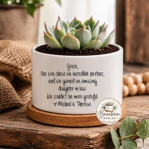 May include: A white ceramic cylindrical planter containing a green succulent plant. The planter features black printed text that reads: Grace, Our son chose an incredible partner, and we gained an amazing daughter-in-law. We couldn't be more grateful. Michael & Theresa.