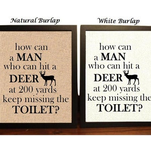May include: Two framed burlap signs with black frames. The signs have the same text: "How can a man who can hit a deer at 200 yards keep missing the toilet?" The sign on the left has a natural burlap background and the sign on the right has a white burlap background.