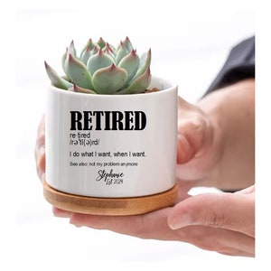 May include: White ceramic planter with the word "RETIRED" in bold black letters. The definition of "retired" is written below, along with the phrase "I do what I want, when I want." The text "See also: not my problem anymore" is written below that. The planter has a bamboo base and a small succulent plant growing inside.