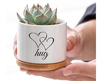 May include: White ceramic succulent planter with a bamboo base. The planter has two intertwined hearts and the word "hug" printed on it.
