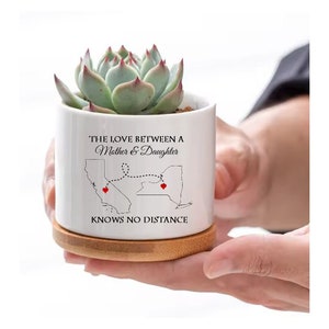 May include: White ceramic succulent planter with a black and white graphic of the states of California and New York connected by a dotted line. The text "The love between a Mother & Daughter knows no distance" is printed on the planter.