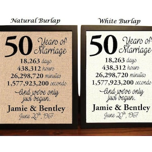 May include: Two framed burlap prints with black frames. The prints are identical except for the background color. The prints feature the text "50 Years of Marriage" with the number 50 in large font. The prints also include the number of days, hours, minutes, and seconds in 50 years. The text "And we've only just begun..." is included below the numbers. The names "Jamie & Bentley" and the date "June 20th, 1967" are at the bottom of the prints. The left print has a natural burlap background and the right print has a white burlap background.