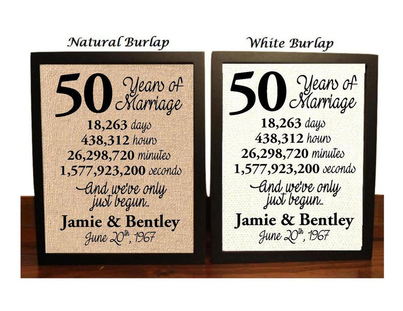 May include: Two framed burlap prints with black frames. The prints are identical except for the background color. The prints feature the text "50 Years of Marriage" with the number 50 in large font. The prints also include the number of days, hours, minutes, and seconds in 50 years. The text "And we've only just begun..." is included below the numbers. The names "Jamie & Bentley" and the date "June 20th, 1967" are at the bottom of the prints. The left print has a natural burlap background and the right print has a white burlap background.