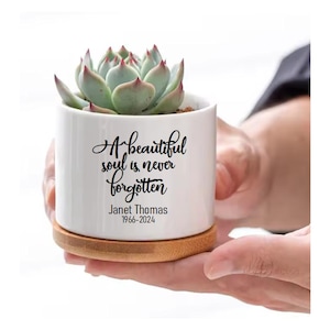 May include: White ceramic planter with a succulent plant. The planter has a black inscription that reads "A beautiful soul is never forgotten" and "Janet Thomas 1966-2024". The planter sits on a wooden base.
