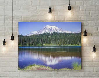 May include: A photograph of a mountain with snow on the peak reflected in a still lake. The mountain is surrounded by a forest of evergreen trees.