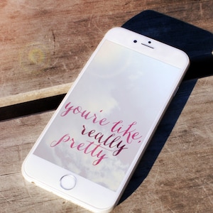 May include: A white smartphone with a pink and white text overlay that reads "you're like, really pretty" on the screen. The phone is lying on a wooden surface.