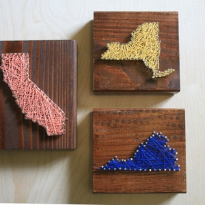 May include: Three wooden plaques with string art depicting the outlines of the states of California, New York, and Virginia. The California outline is pink, New York is gold, and Virginia is blue. The wood has a dark brown stain.