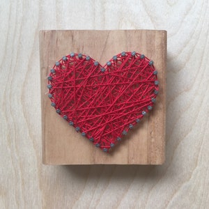 May include: A handmade string art piece featuring a red heart design on a square wooden block. The heart is formed with red thread woven between small nails hammered into the wood. The wood has a natural, light brown finish.