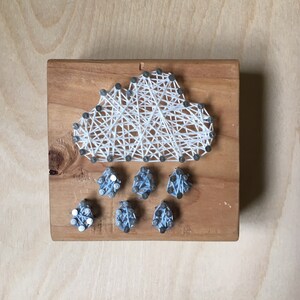May include: A handmade string art piece depicting a cloud with rain. The cloud is crafted with white string on a wooden block, with silver nails outlining the shape. Raindrops are represented by small blue string bundles.