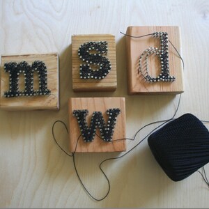 May include: Five wooden blocks with the letters "m", "s", "d", "w", and a spool of black thread. Each letter is made of nails hammered into the wood and strung with black thread.