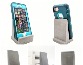 May include: A set of four gray concrete phone stands. Two stands are shown with phones in them, one with a blue phone case and one with a black and blue phone case. The other two stands are shown separately. The stands are designed to hold a phone upright.