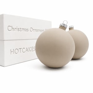 May include: Two matte beige spherical Christmas ornaments with silver-toned metal caps. The ornaments are positioned next to a white rectangular box featuring the embossed text "Christmas Ornament" and "HOTCAKES" on the side of the packaging.