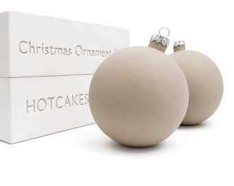 May include: Two matte beige spherical Christmas ornaments with silver-toned metal caps. The ornaments are positioned next to a white rectangular box featuring the embossed text "Christmas Ornament" and "HOTCAKES" on the side of the packaging.