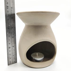 May include: A beige ceramic oil burner with a round opening for a tea light candle. The burner has a wide, flat top and a rounded bottom.