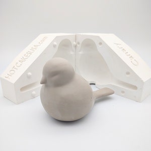 May include: A white two-piece mold for making a bird figurine. The mold is open and a gray bird figurine is sitting in front of it. The mold is labeled "HOTCAKESUSA.com" and "Candy".