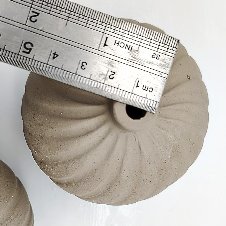 May include: Two small, brown, ceramic planters with a fluted design. The planters are approximately 2 inches in diameter. A ruler is used to show the size of the planters.