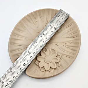 May include: A light brown ceramic dish with a raised flower design in the center. The dish is approximately 6 inches in diameter.