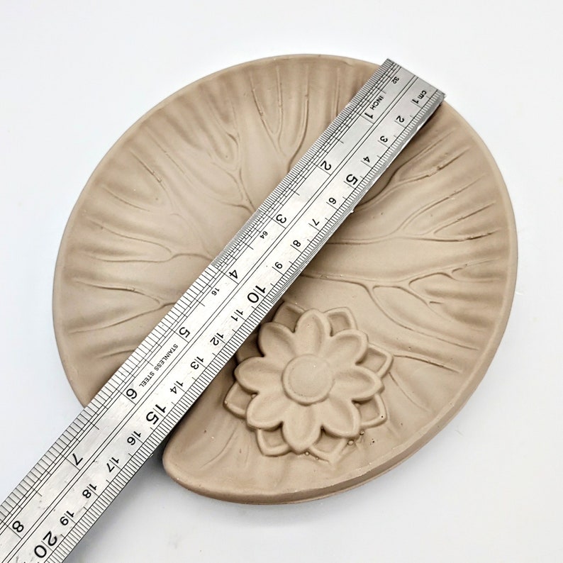 May include: A light brown ceramic dish with a raised flower design in the center. The dish is approximately 6 inches in diameter.