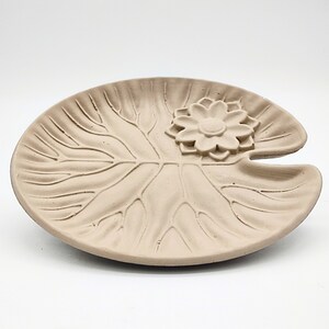 May include: A light brown ceramic dish shaped like a lily pad with a raised flower design in the center.