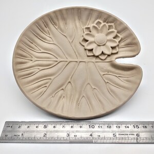 May include: A light brown ceramic dish shaped like a lily pad with a raised flower design in the center. The dish is approximately 5 inches in diameter.