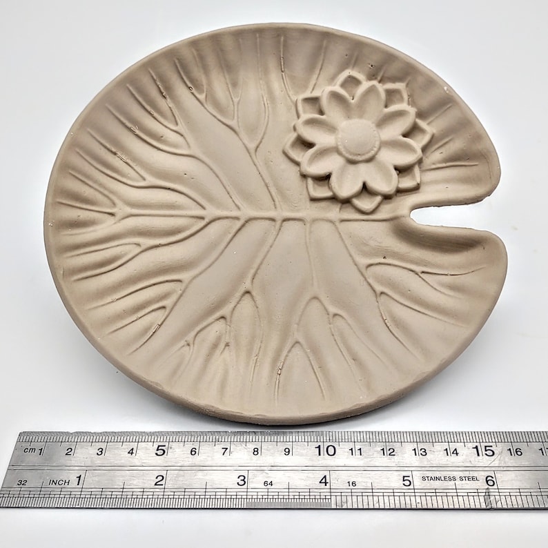 May include: A light brown ceramic dish shaped like a lily pad with a raised flower design in the center. The dish is approximately 5 inches in diameter.
