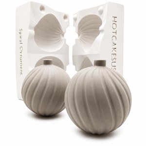 May include: Two light gray spherical ornaments with a spiral ribbed texture and a small neck, positioned in front of a white two-part plaster mold. The mold features the embossed text Spiral Ornament and HOTCAKESUS on its vertical sides.
