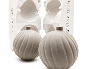 May include: Two light gray spherical ornaments with a spiral ribbed texture and a small neck, positioned in front of a white two-part plaster mold. The mold features the embossed text Spiral Ornament and HOTCAKESUS on its vertical sides.
