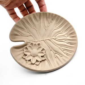 May include: A light brown ceramic dish shaped like a lily pad with a raised flower design in the center.