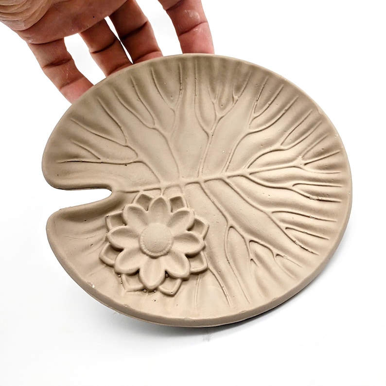 May include: A light brown ceramic dish shaped like a lily pad with a raised flower design in the center.