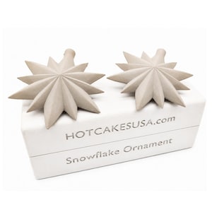 May include: Two light beige, multi-pointed star-shaped ornaments resting on a white rectangular box. The box features the text HOTCAKESUSA.com and Snowflake Ornament printed in a metallic silver font on the front side of the packaging.