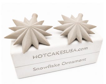 May include: Two light beige, multi-pointed star-shaped ornaments resting on a white rectangular box. The box features the text HOTCAKESUSA.com and Snowflake Ornament printed in a metallic silver font on the front side of the packaging.