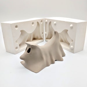 May include: Two white plaster molds for casting a ceramic ghost figurine. The molds are open and the ceramic ghost is sitting in between them.
