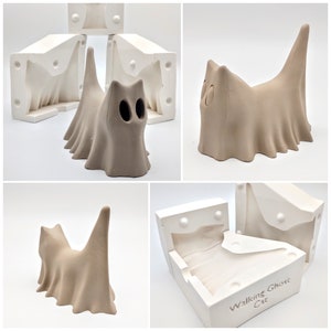May include: A white plaster mold for making a ceramic sculpture of a cat wearing a ghost costume. The mold is in two pieces and has the words "Walking Ghost Cat" written on it.