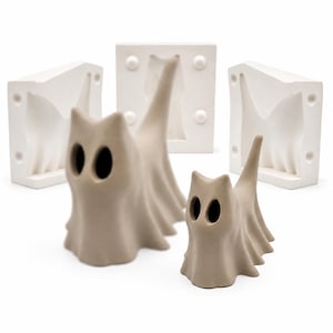May include: Two beige, ghost-like cat figurines with hollow oval eyes and draped bodies, positioned in front of three white, rectangular plaster molds that feature recessed cat-shaped cavities and circular registration points.