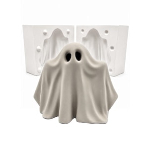 May include: A light gray, matte-finished ghost figurine with two dark, oval-shaped eye openings, positioned in front of a two-part white plaster mold. The mold features a hollow, ghost-shaped cavity and circular registration keys on the sides.