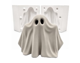 May include: A light gray, matte-finished ghost figurine with two dark, oval-shaped eye openings, positioned in front of a two-part white plaster mold. The mold features a hollow, ghost-shaped cavity and circular registration keys on the sides.