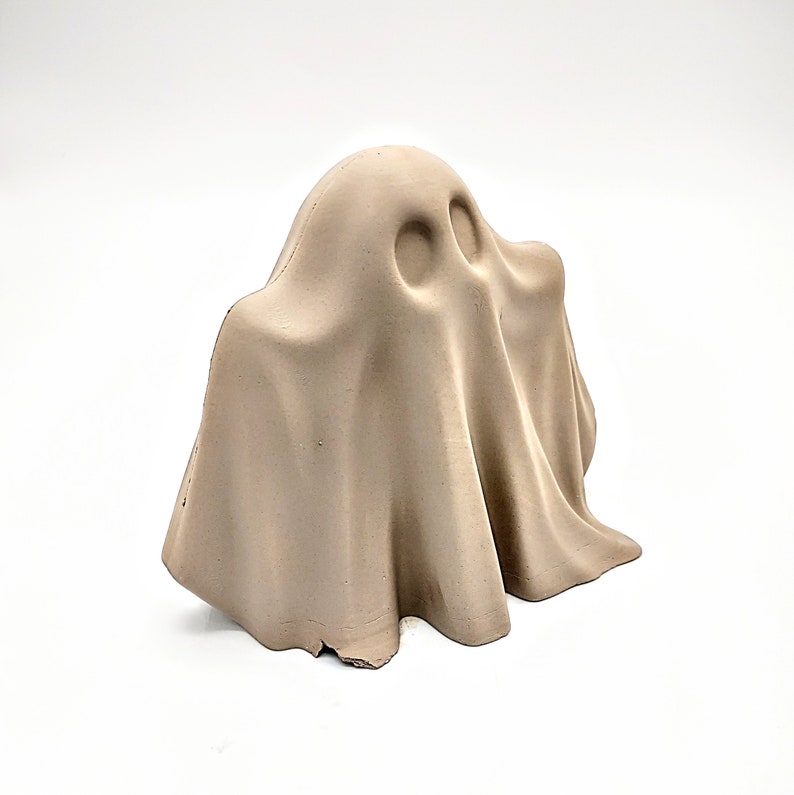 May include: A beige ceramic ghost figurine with a simple, rounded shape. The ghost has a smooth, matte finish and a slightly textured surface.