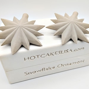 May include: Two light gray snowflake ornaments with pointed edges rest on a white box. The box has the text "HOTCAKESUSA.com" and "Snowflake Ornament" engraved on it. The ornaments are a starburst design.