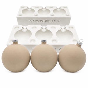 May include: Three matte beige spherical ornaments with silver-toned metal caps positioned in front of a white rectangular mold tray. The mold features three circular indentations and the text HOTCAKESUSA.COM embossed on the front side.