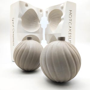 May include: Two gray ceramic ornaments with a spiral design, sitting next to a white mold with the text "HOTCAKESUS" and "Spiral Ornament" on it.
