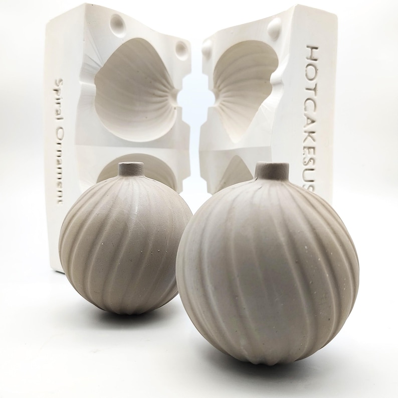 May include: Two gray ceramic ornaments with a spiral design, sitting next to a white mold with the text "HOTCAKESUS" and "Spiral Ornament" on it.